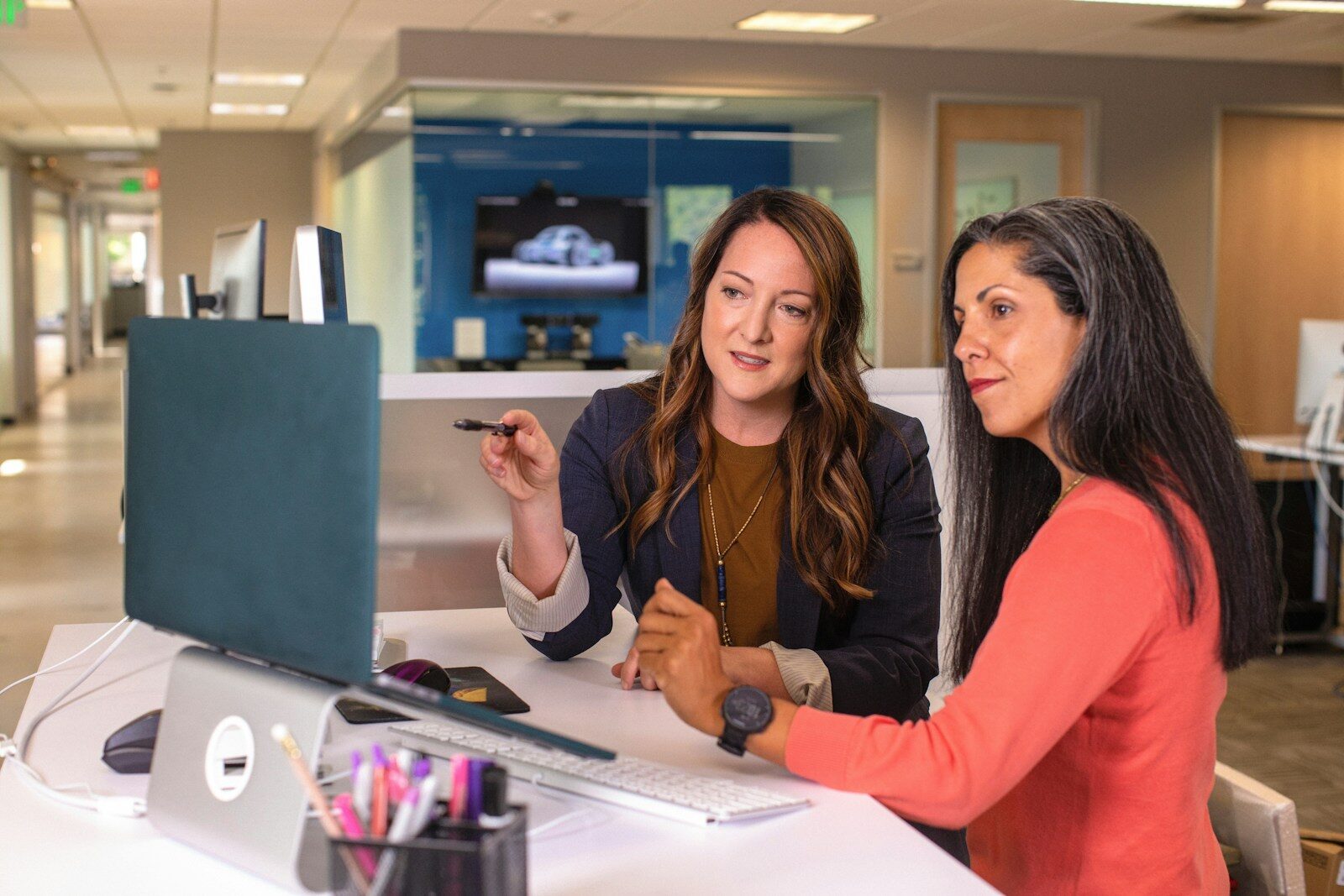 Two women collaborate at a modern office desk, discussing work on a computer. One points to the screen while the other listens attentively.