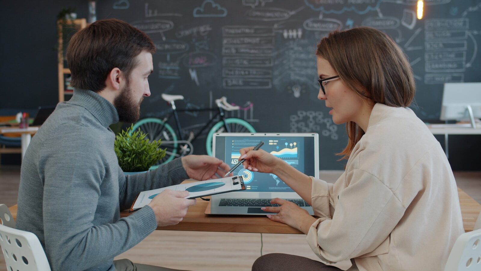 A man and woman collaborate at a desk, discussing data displayed on a laptop while reviewing a document, in a modern office setting.