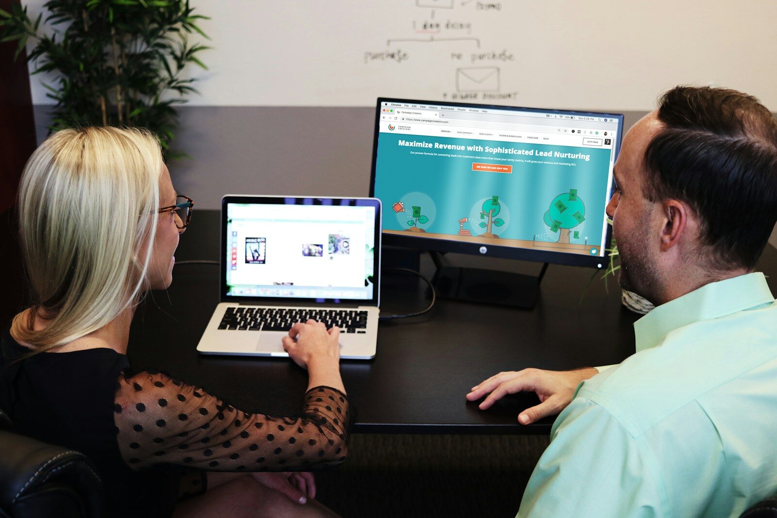 A woman and a man discuss marketing strategies, with the woman using a laptop and the man observing content on a large monitor.