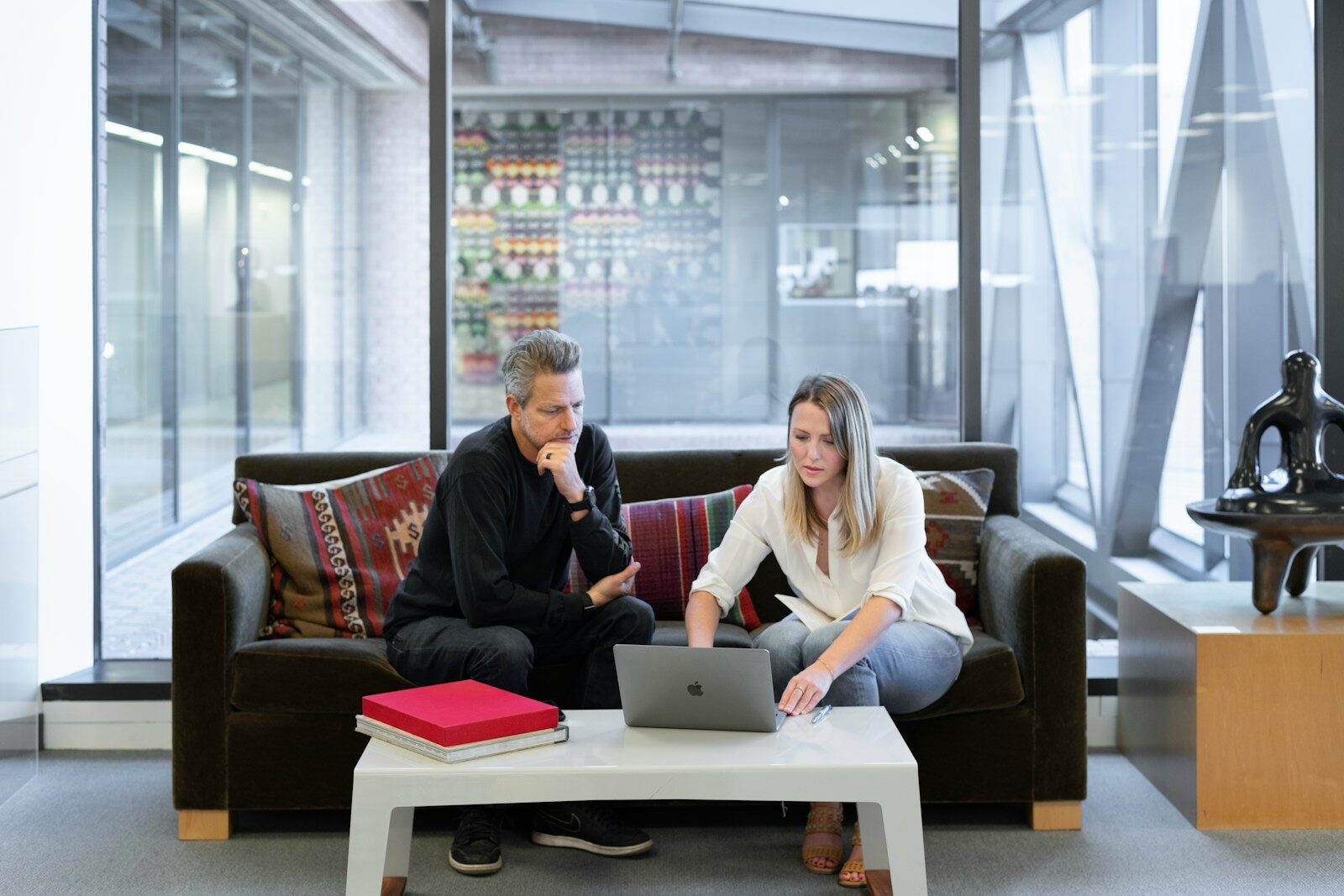 A man and a woman collaborate on a laptop in a modern office setting, discussing work while sitting on a couch with colorful cushions.